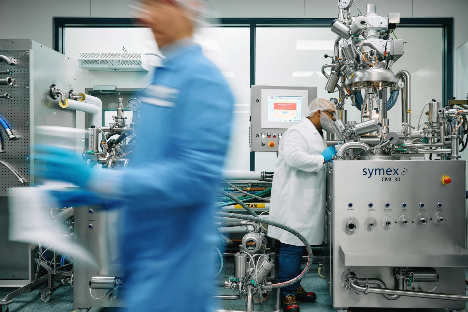 A person in a blue lab coat moving past formulation equipment in a lab.